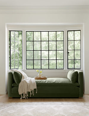 The Elvie moss green velvet chaise sits in a nook with three black-framed windows with a cork tray and ivory throw atop it
