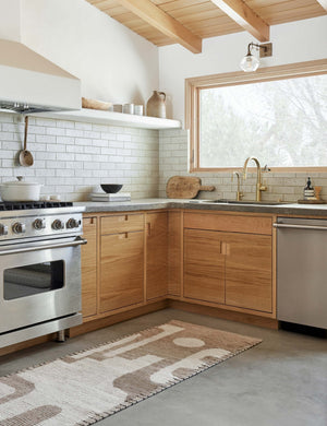 The Abode geometric two-toned kand-knotted floor rug by Élan Byrd with woven border in its runner size sits in a kitchen with wooden cabinetry and a gray subway-tile backsplash atop a concrete floor