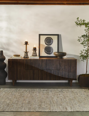 The Lemieux et Cie Pasteur matte black geometric curved Floor Vase sits in a room with a wooden textured sideboard, a textured brown carpet, and a geometric wall art