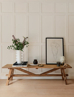 The Arlene craftsman-style antiqued teak wood bench sits against a white paneled wall and has a sketch of a person on it along with decor accessories and a white vase full of stems.