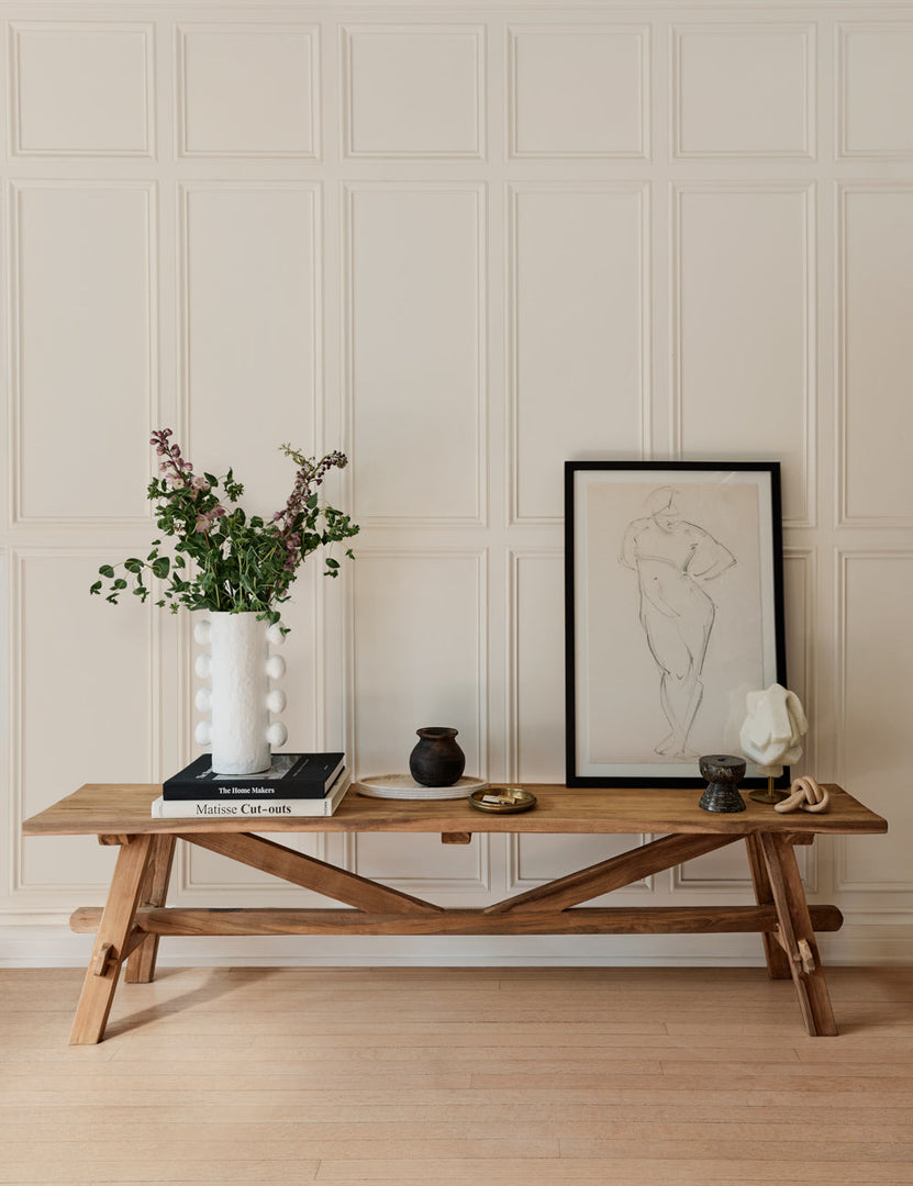 | The Arlene craftsman-style antiqued teak wood bench sits against a white paneled wall and has a sketch of a person on it along with decor accessories and a white vase full of stems.