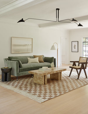 The Brisa rectangular burl wood coffee table with four legs sits in a modern living room with a moss green sofa, a tan and ivory checked rug, and a brown wood and cane accent chair under a large modern black chandelier.