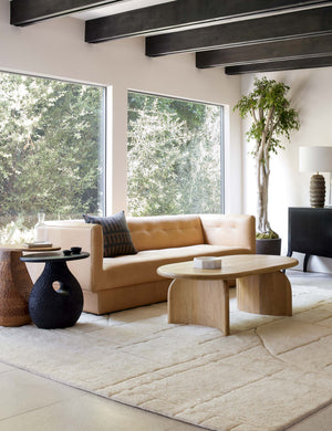 The Ada solid oak oval coffee table sits in an open living room with a light tan leather sofa and ivory area rug. Dark stained wood beams line the ceiling.