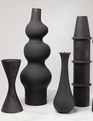 The Lemieux et Cie Pasteur matte black geometric curved Floor Vase sits in a room with other matte black geometric floor vases