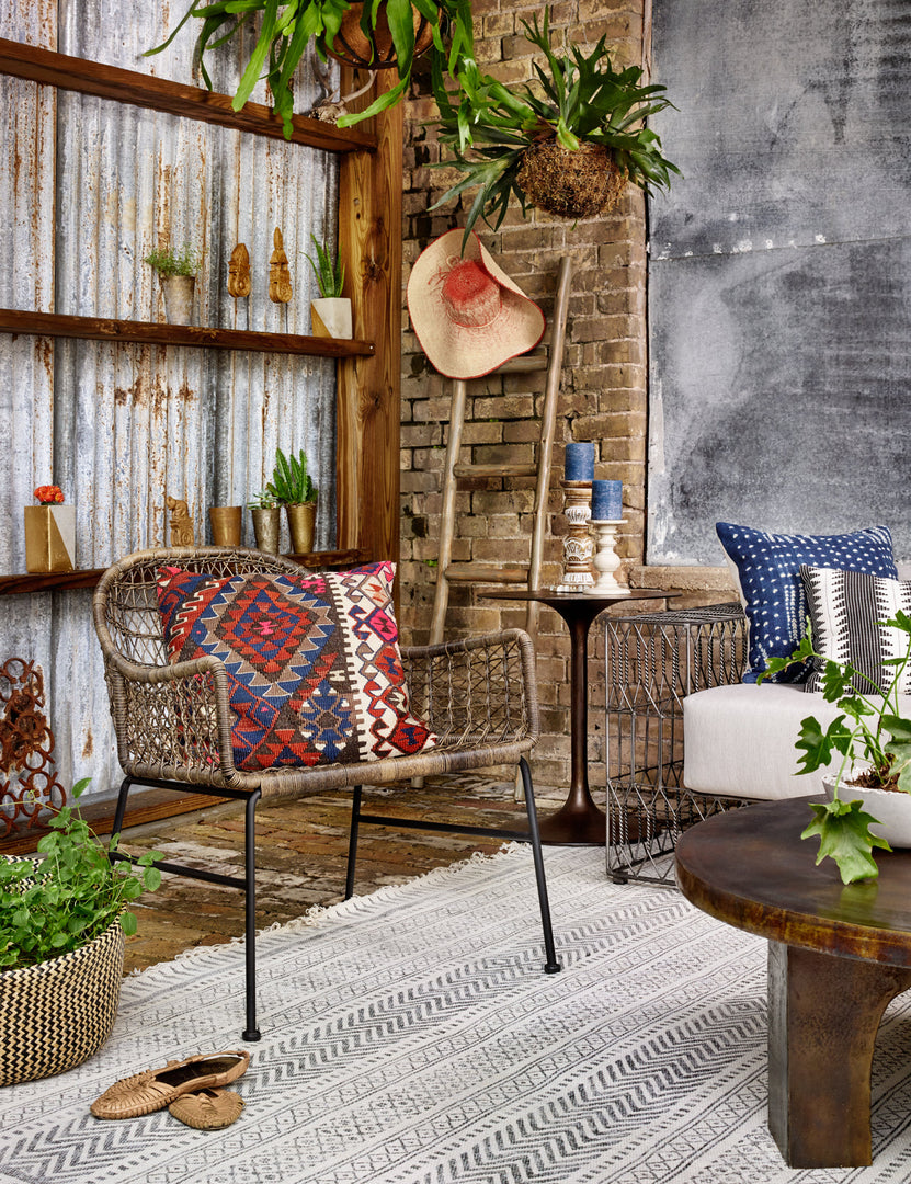 | The Eliza brown open-woven wicker indoor and outdoor accent chair sits in an industrial living room with a brick accent wall, a blue and white patterned rug, and a colorful throw pillow sitting atop it