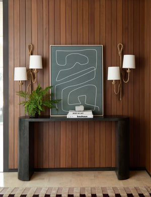Two Coastal living bimini woven rattan double sconces by Regina Andrew are hung around a gray patterned wall art above a white wooden sideboard against a wood paneled wall