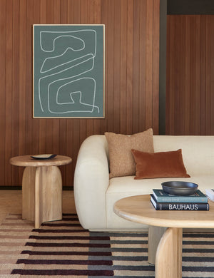 The Ada solid oak side table with oval top and curved pedestal base sits in a dark paneled living room with a curved ivory sofa with brown throw pillows and a matching Ada oak coffee table.