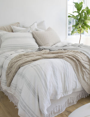 The Jackson Linen white and ocean striped Sham by Pom Pom at Home lays on a bed in a bedroom with textured throw pillows, bright windows, and hardwood floors